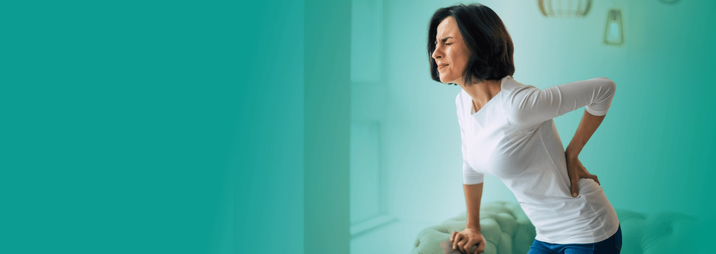 A woman touching her lower back while trying to stand from a couch and feeling pain.