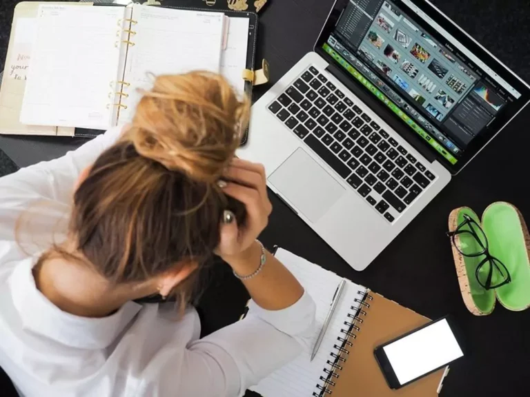 A stressed woman in front of a laptop and a pile of paperwork.